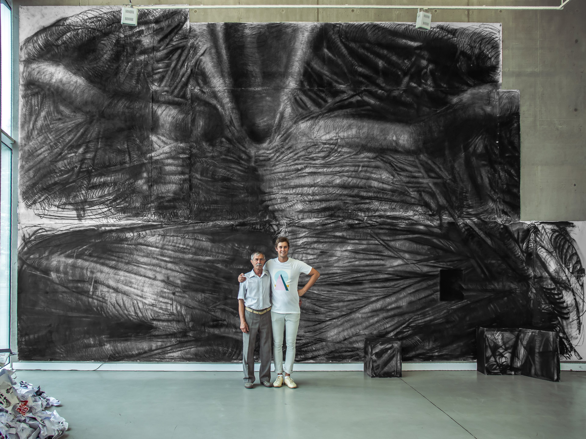 Vasyl Savchenko holds his grandfather by the shoulder in front of the large scale, detailed charcoal drawing of an anthropomorphic form hanging on the wall